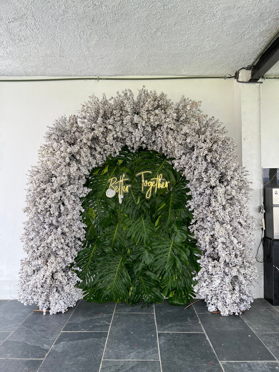 Arco floral de bienvenida de flor blanca nevada, con muro verde y letrero de neón, en una terraza