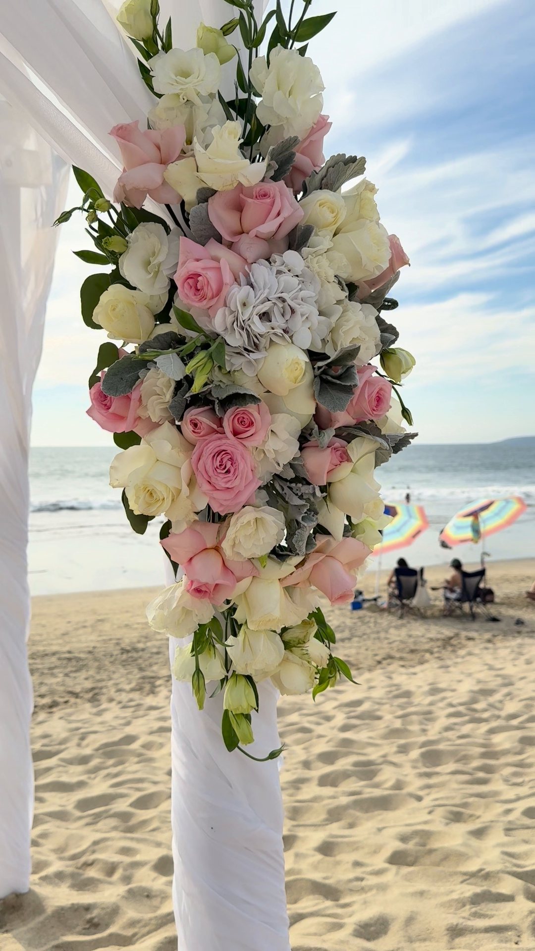 Ramo de flor rosa y blanca con follaje en la esquina de un arco de ceremonia con telas, en la playa