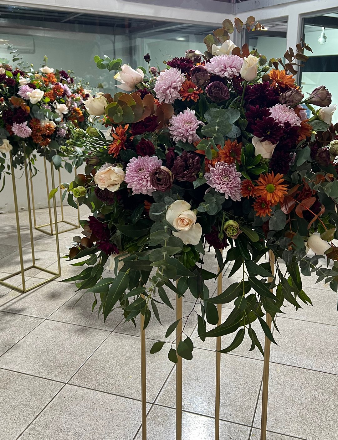 Centro de mesa alto de flor otoñal en rosa, vino y naranja sobre base dorada