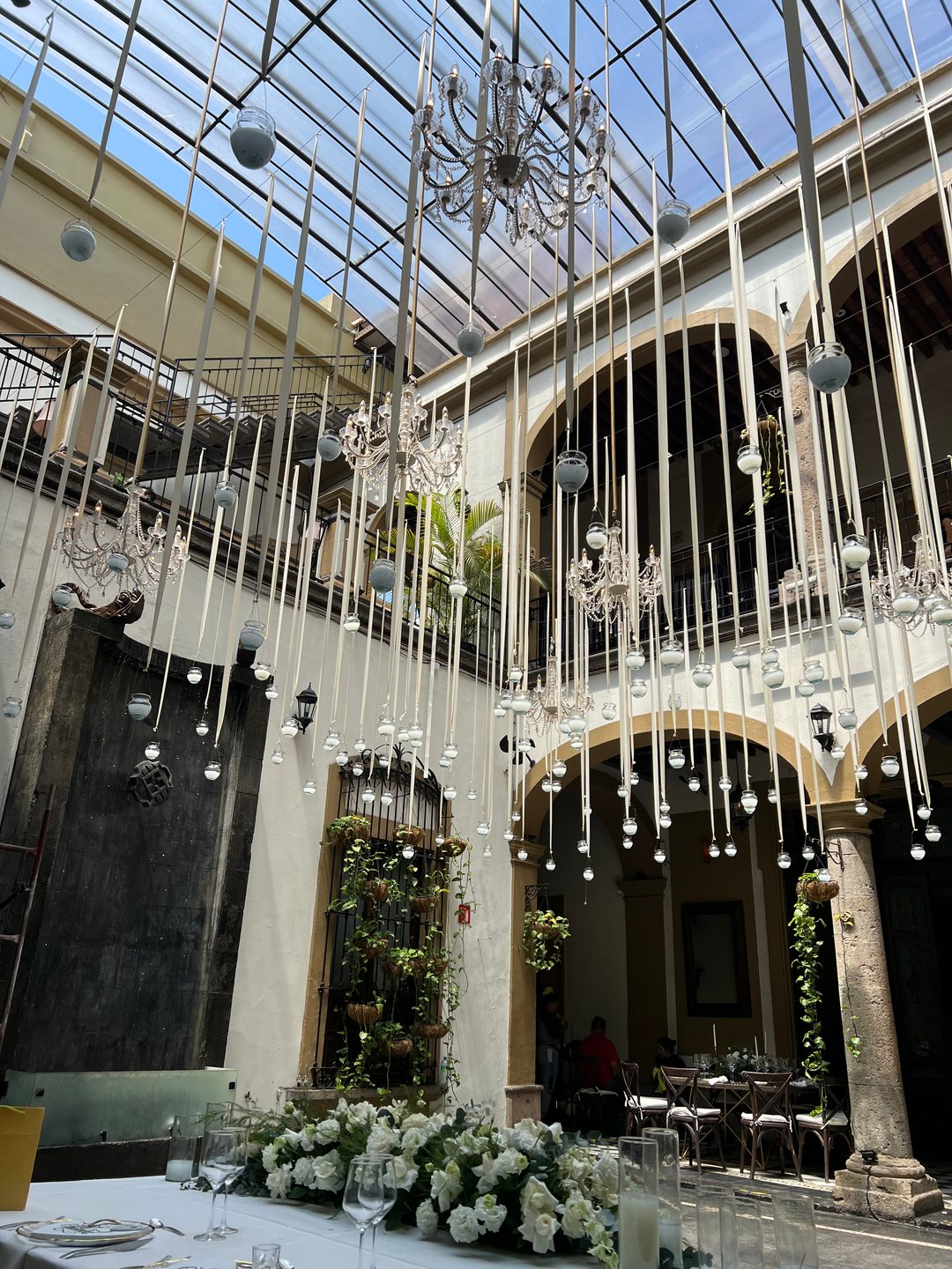Arreglos colgantes: listones blancos con veladoras y candiles de cristal bajo el techo de vidrio de un patio, Guadalajara