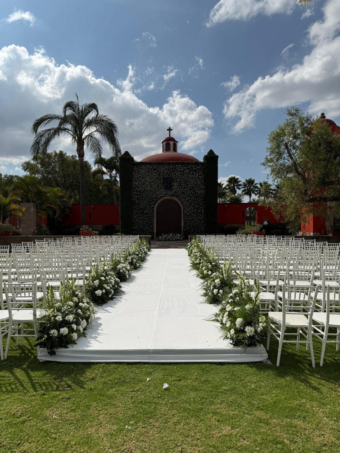 Pasillo de la ceremonia con flor blanca en las sillas y la capilla forrada de follaje al fondo, Zapopan