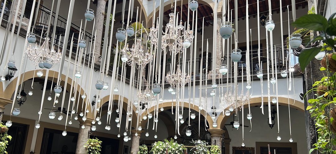 Veladoras colgantes y listones blancos con candiles en un patio colonial con arcos y muros de follaje, Guadalajara