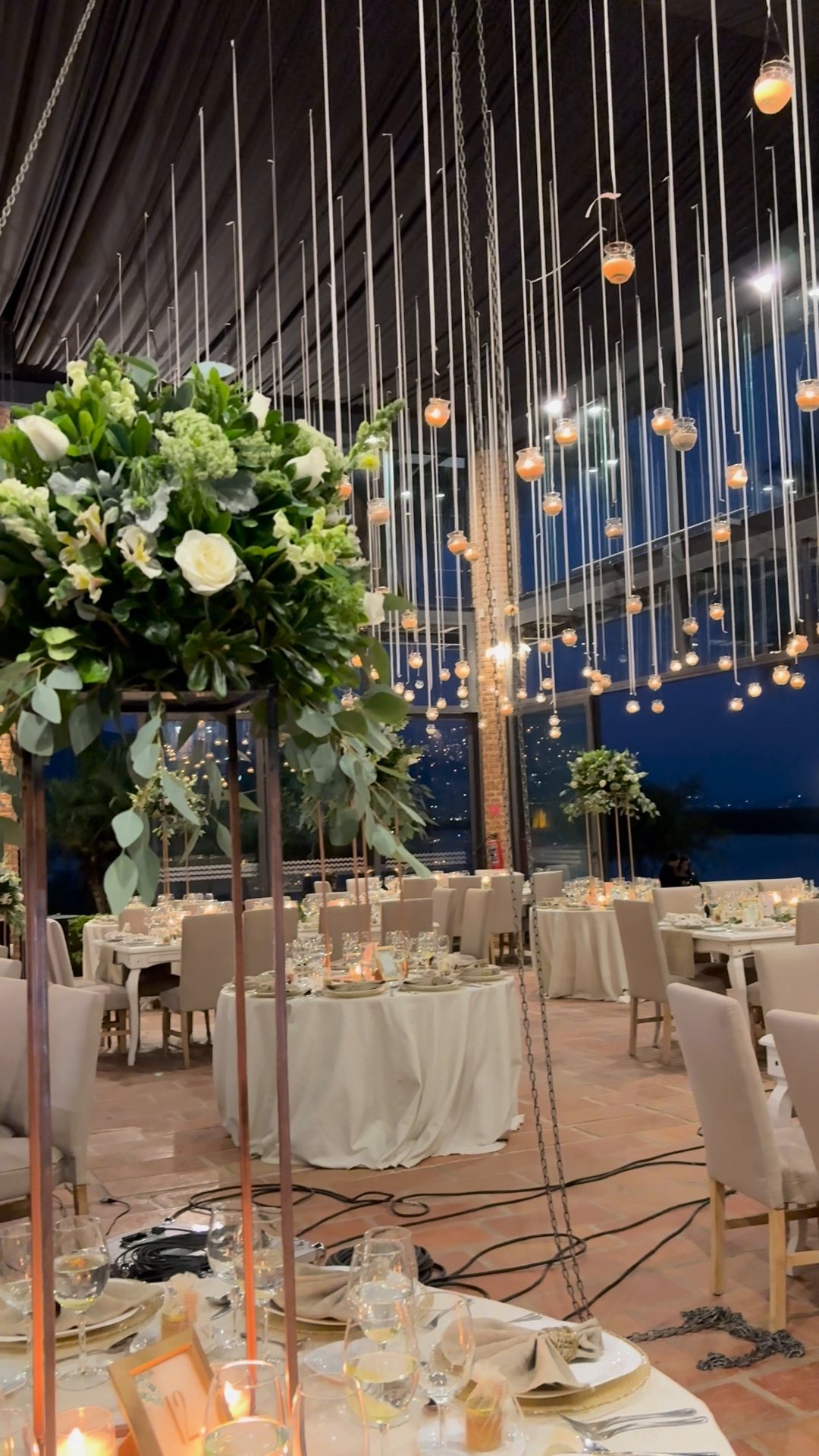 Centro de mesa alto de follaje y flor blanca bajo series de focos colgantes, salón de cristal de noche