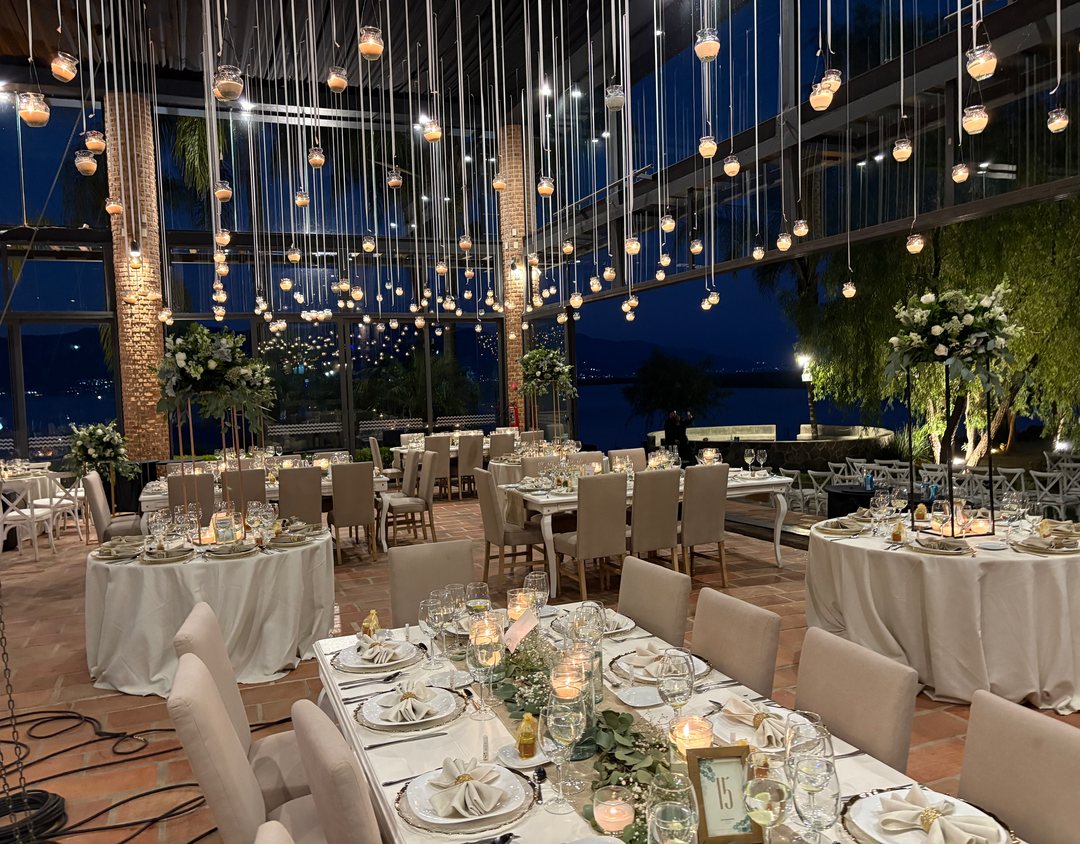 Salón de cristal de noche junto al lago, con cientos de veladoras colgando de listones, centros altos de flor blanca y caminos de follaje con velas en las mesas