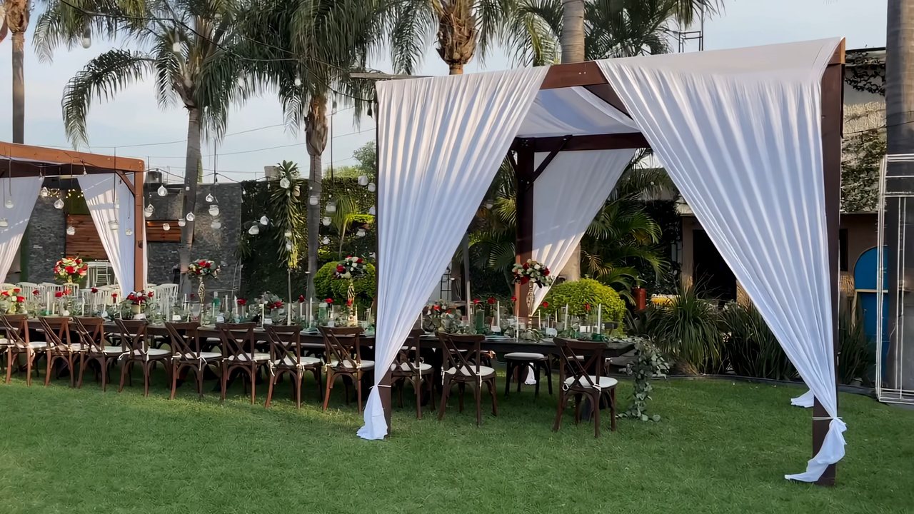 Pérgolas enteladas con cortinas blancas que se mecen sobre una mesa imperial con flor roja y blanca, de día