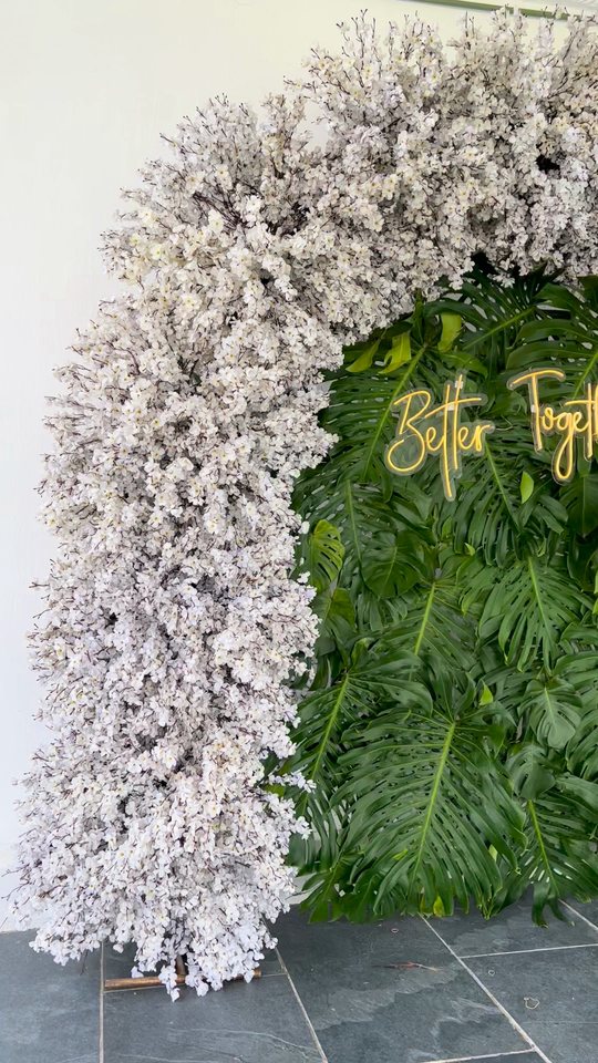 Arco de flor blanca nevada alrededor de un muro de follaje verde con letrero de neón