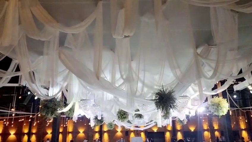 Techo drapeado de tela blanca con candiles de cristal y esferas de follaje colgantes, de noche