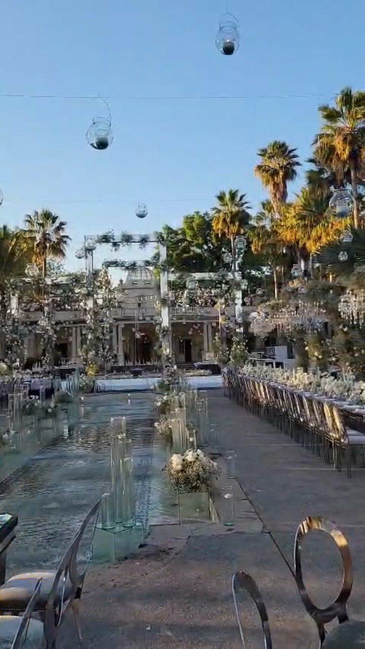 Recorrido por un jardín con palmeras, mesas imperiales con flor blanca y esferas de vidrio con velas colgando, al atardecer
