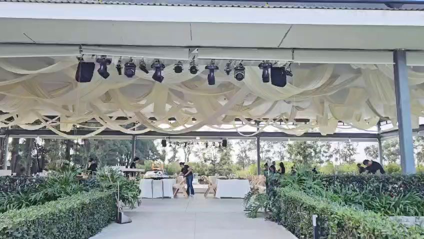 Ondas de tela crema con luces robóticas sobre una terraza con jardín, mientras se montan las mesas