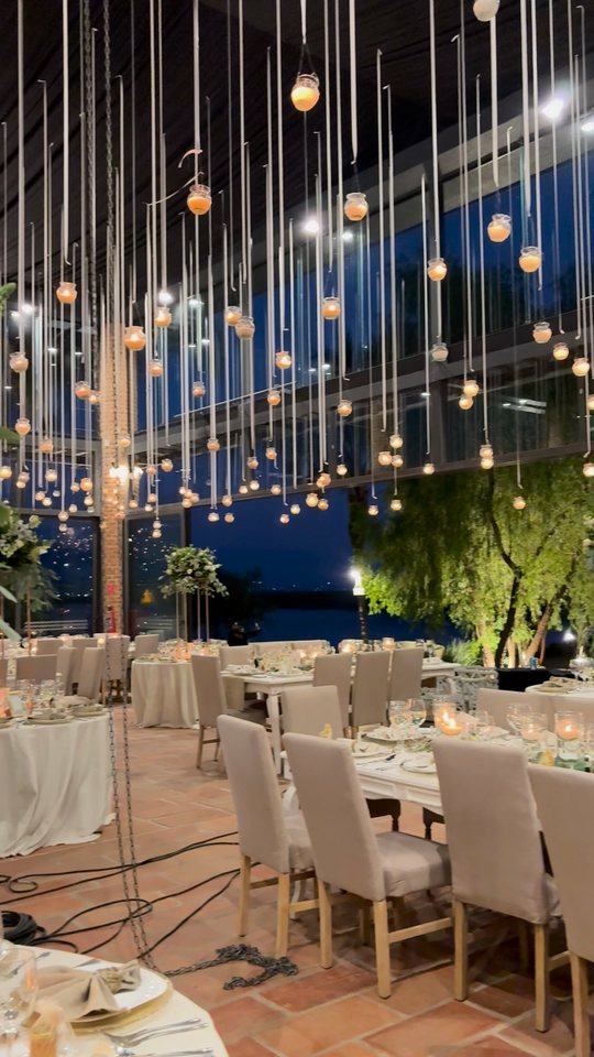 Recorrido de noche por un salón de cristal con veladoras colgantes, mesas montadas y un centro alto de follaje y flor blanca