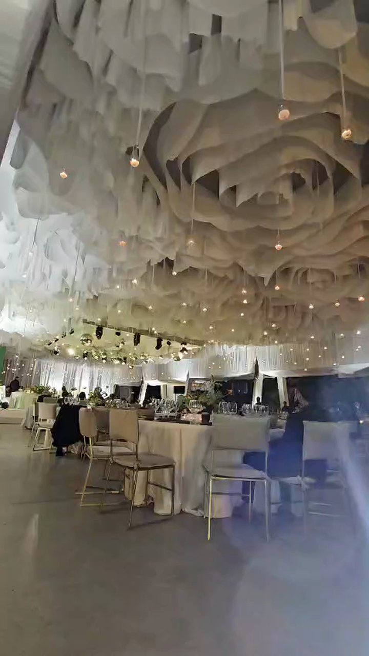 Techo de tela blanca en pliegues tipo rosa con velas colgantes en globos de cristal sobre un salón con mesas, de noche
